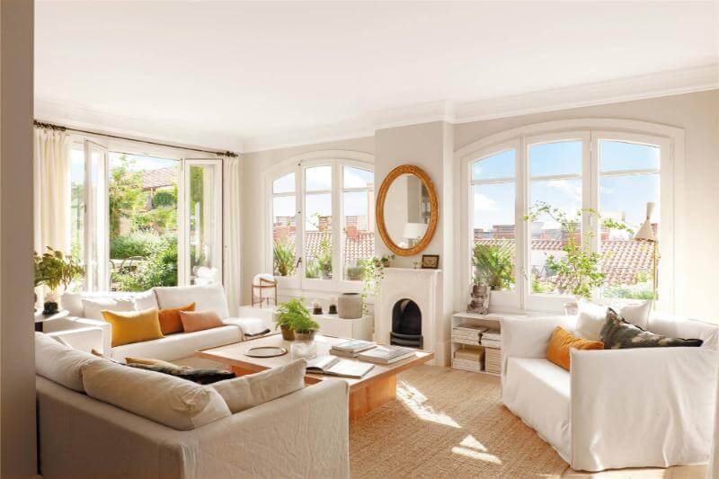Una sala de estar con grandes ventanas, cortinas blancas y un espejo grande reflejando la luz del sol. Preparar nuestra casa para el verano