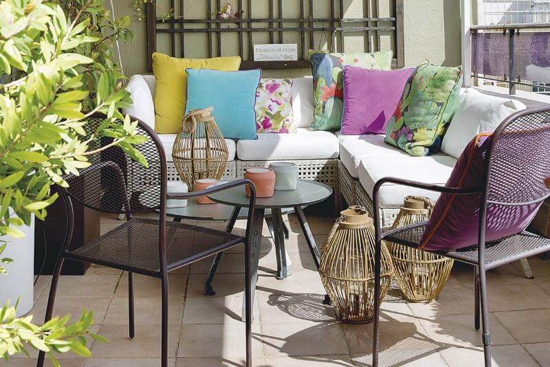 Terraza decorada para Vivir el Verano al Máximo con un sofá de exterior blanco y cojines multicolores, acompañada de una mesa de centro y sillas de metal, rodeada de plantas.