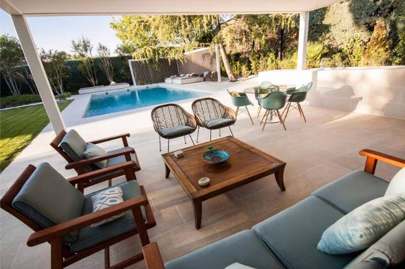 Área de piscina al aire libre con sofás y sillas, rodeada de plantas y césped con una vista despejada del agua. Para Vivir el Verano al Máximo.