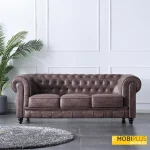 Sofá Chesterfield marrón chocolate en salón moderno con molduras blancas y planta de palmera, de MobiPlus Muebles.